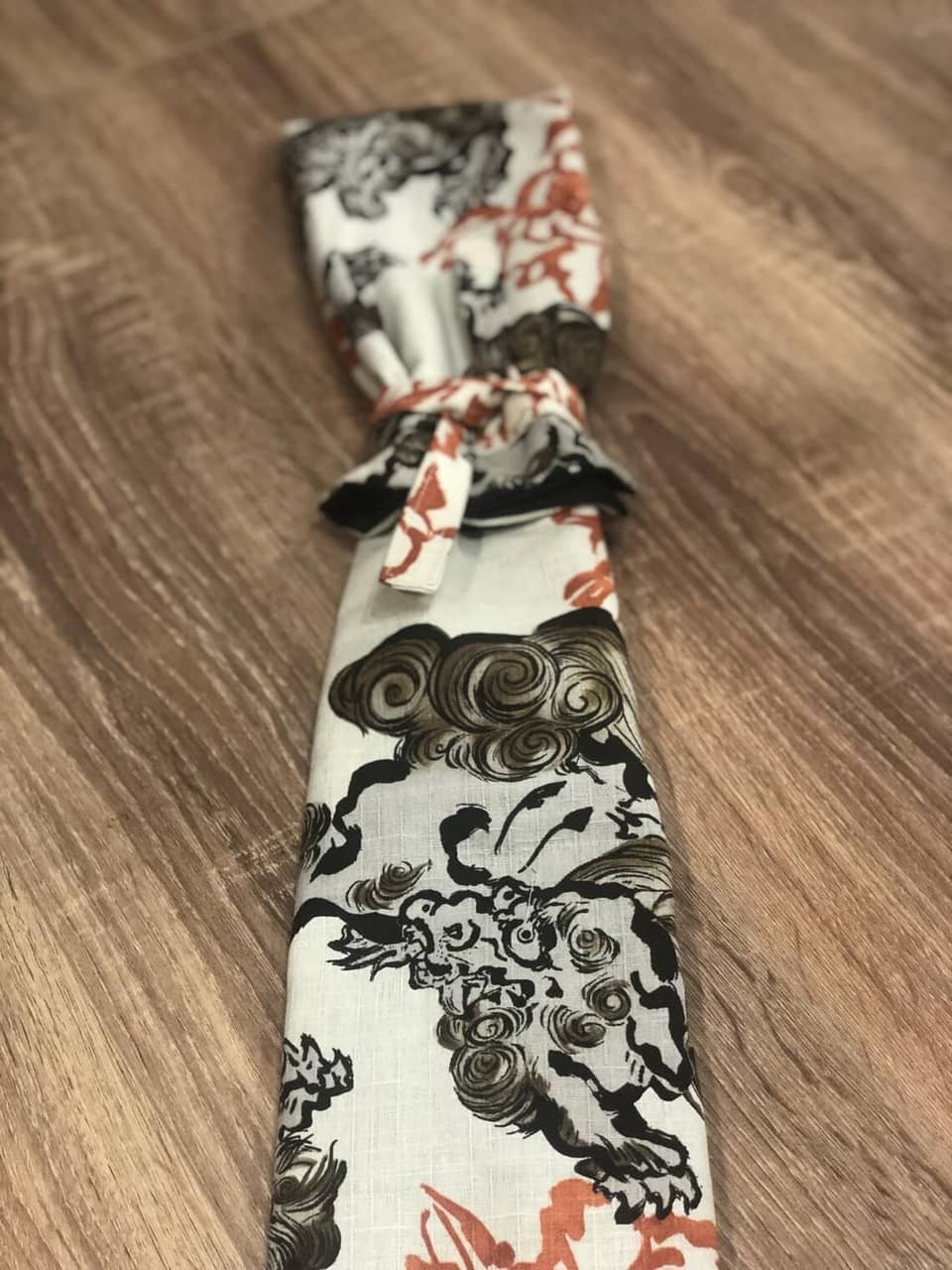 Una bolsa de espada de tela blanca con un diseño marrón y naranja de una criatura mítica. La bolsa está atada con una cinta marrón.
