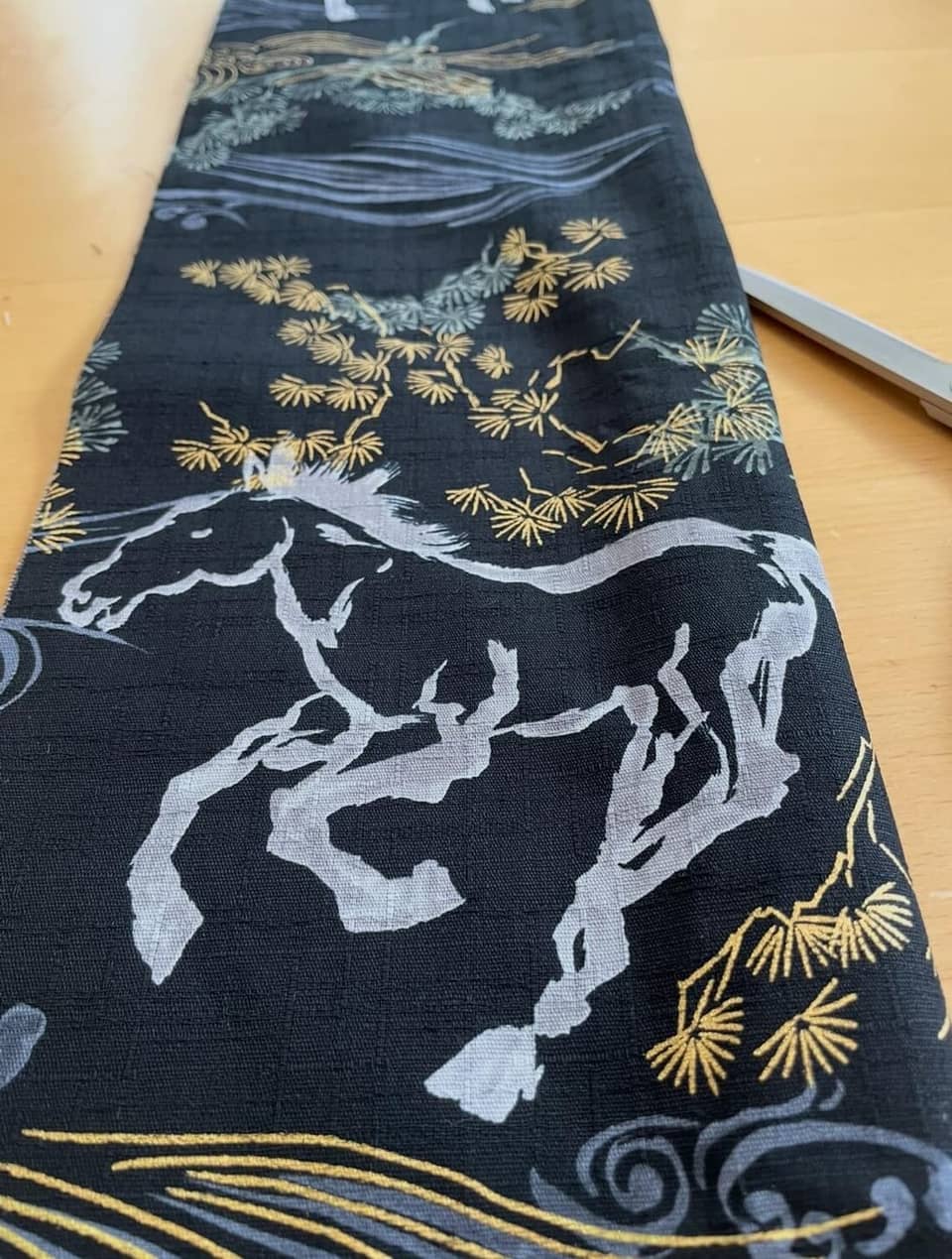 Tela negra con un diseño de caballo corriendo en gris y blanco. La tela también presenta ramas de pino doradas y grises y patrones de olas. La tela es probablemente para coser o manualidades.