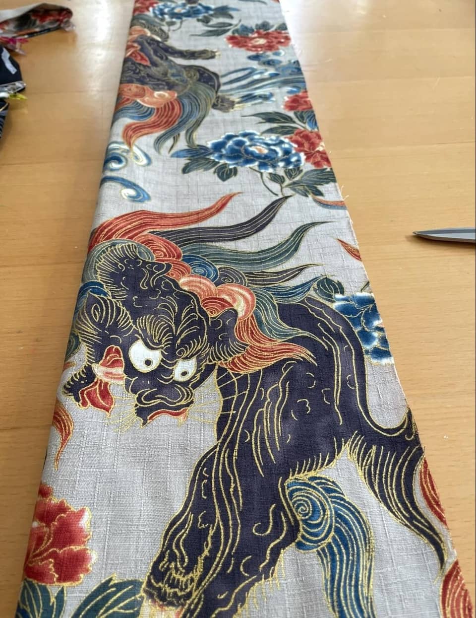 Una tira larga de tela gris claro con un patrón repetitivo de criaturas míticas azul oscuro con contornos dorados, intercaladas con motivos florales rojos y azules. El tejido tiene una textura visible y un diseño detallado.