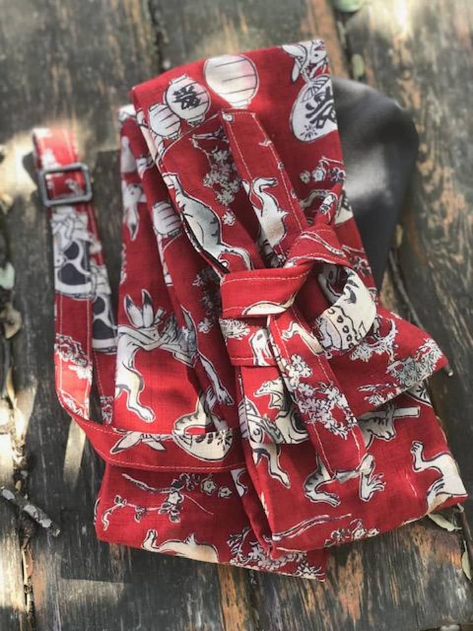 Bolso de tela roja con correa y hebilla, con un patrón repetido de ilustraciones blancas y grises de animales, linternas y caracteres japoneses. El bolso está doblado y atado, con un elemento de cuero negro visible.