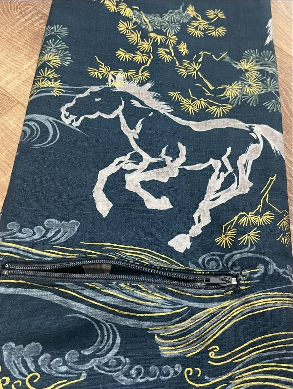 Un estuche de tela color verde azulado oscuro con un diseño estampado de un caballo blanco galopando sobre un paisaje con pinos amarillos estilizados y patrones de agua en gris y dorado. El estuche incluye una cremallera negra horizontal.