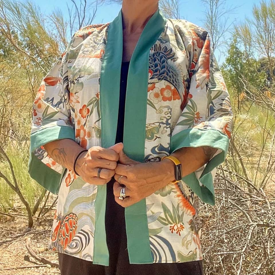 Una chaqueta tipo kimono ligera abierta por delante, con fondo crema y un estampado multicolor de grullas, tortugas y motivos florales. La prenda presenta un borde de color verde azulado sólido a lo largo de la abertura frontal.