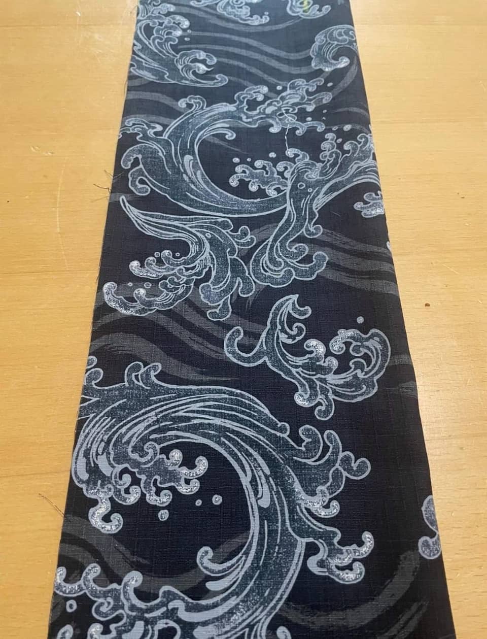 Una tira rectangular de tela negra con un patrón repetitivo de olas oceánicas estilizadas en azul claro y blanco. El diseño incluye corrientes de agua arremolinadas y detalles de espuma impresos sobre el fondo oscuro.